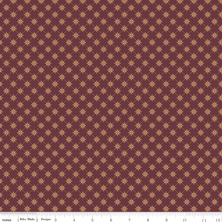 A close-up fabric pattern swatch showing a repeating geometric design of small tan starburst motifs on a muted burgundy background, with a ruler along the bottom edge for scale.