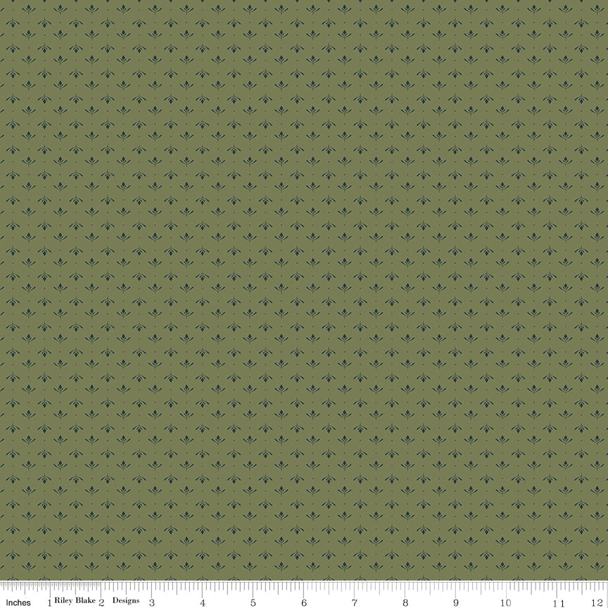 A close-up fabric pattern swatch showing an olive green background with a repeating small dark blue fleur-de-lis motif, with a ruler along the bottom edge for scale.
