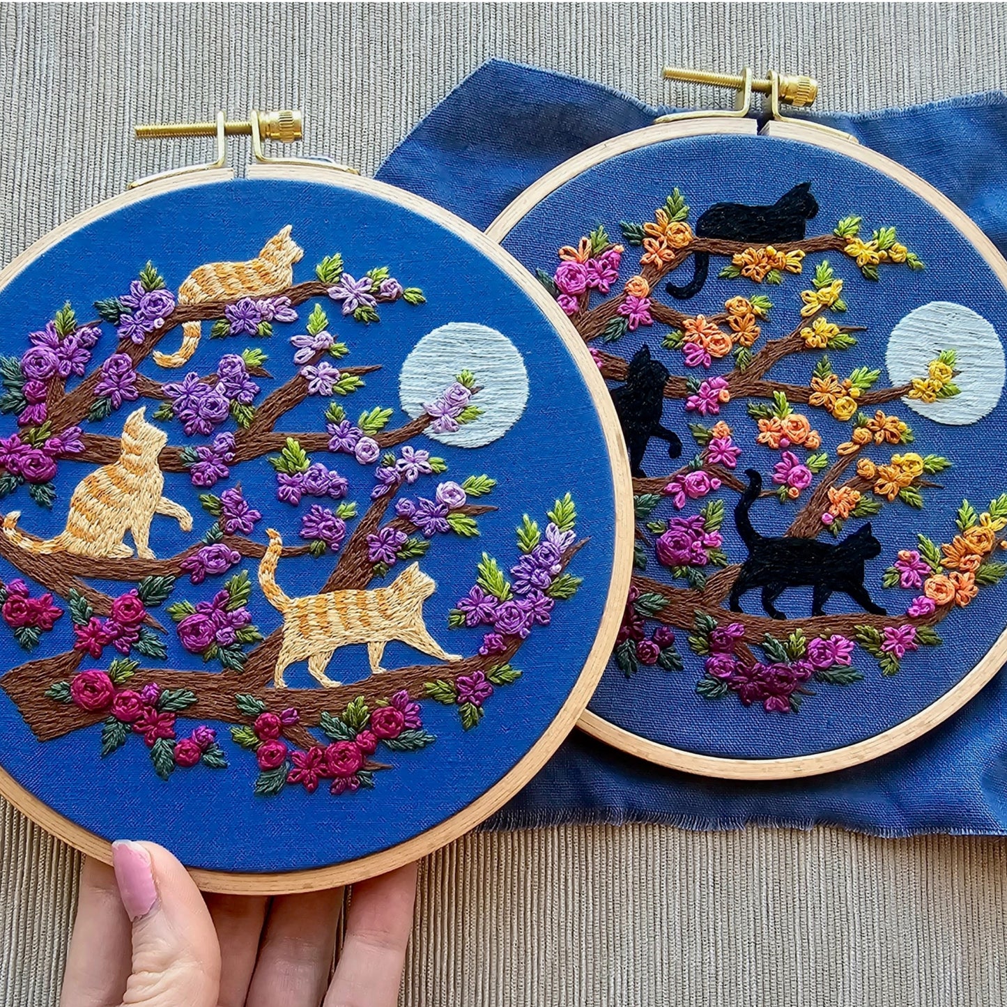 Two embroidery hoops with cat-themed designs on a blue fabric background.