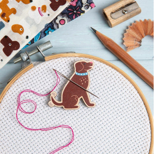 A wooden dog-shaped needle minder with white Aida fabric, pink thread, and a needle, styled on a light blue wooden surface with a pencil, pencil shavings, and patterned fabric.