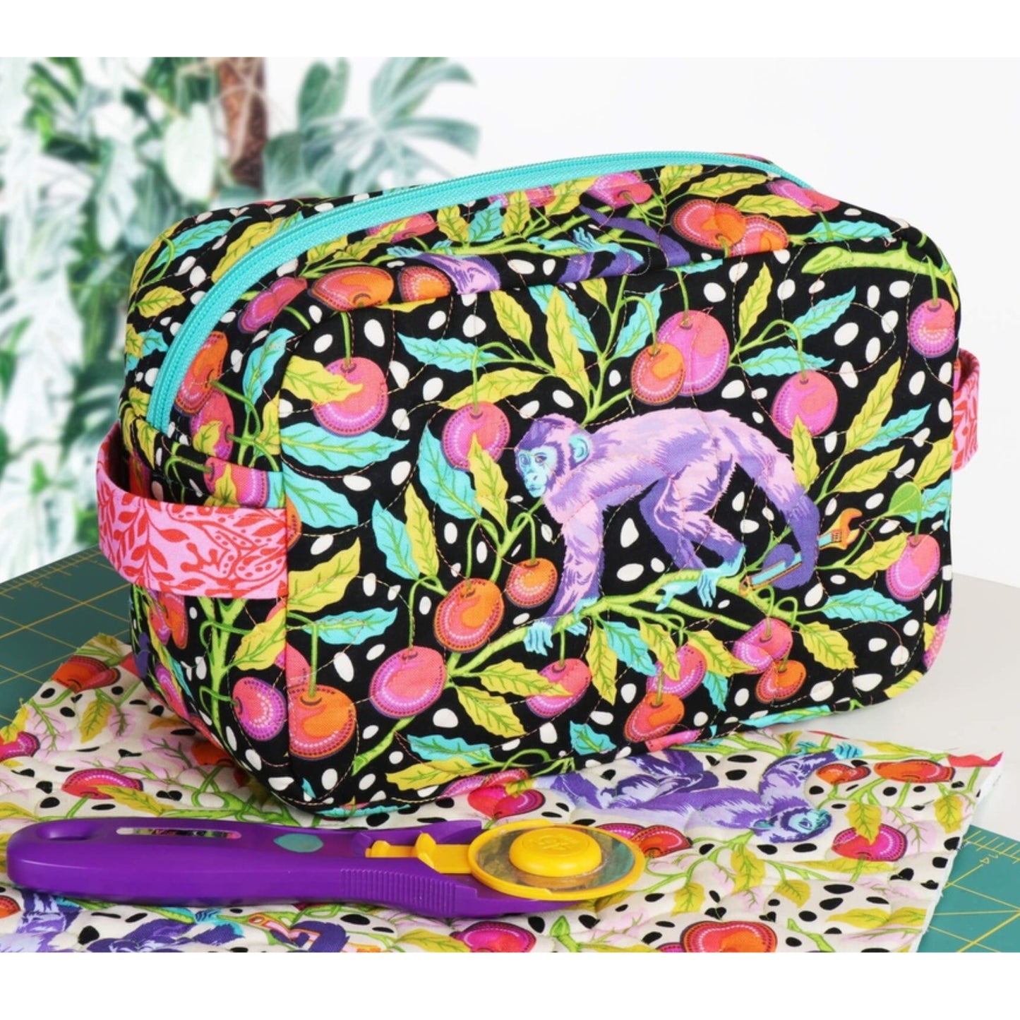A colorful zippered fabric pouch with a black background, white polka dots, and a vibrant tropical fruit and leaf print, featuring a purple sloth graphic. The pouch is styled on a table with a purple rotary cutter and a matching fabric swatch.