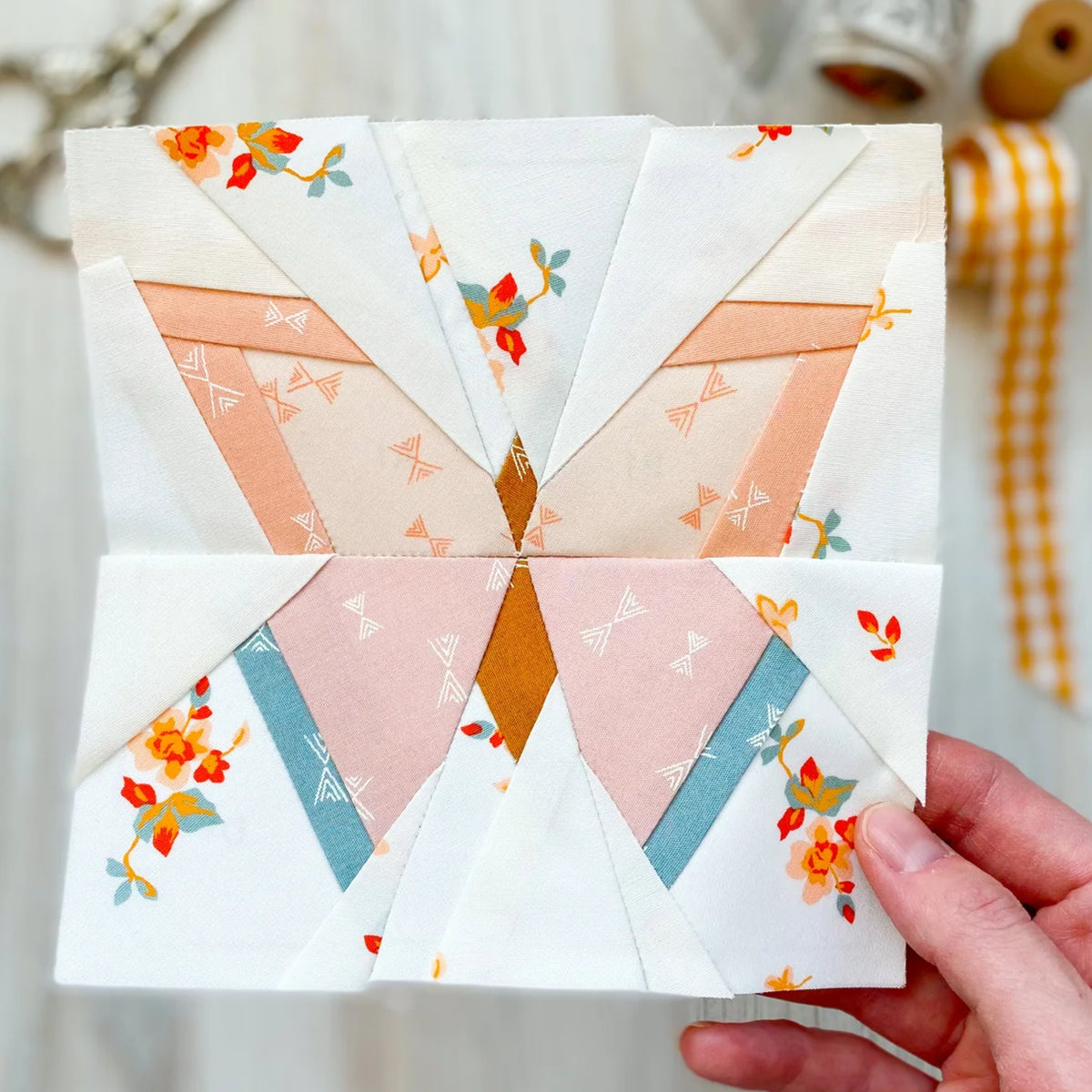 A hand holding a square quilt block made from patterned fabric triangles in soft peach, pink, blue, orange, and white, with a blurred craft workspace in the background.