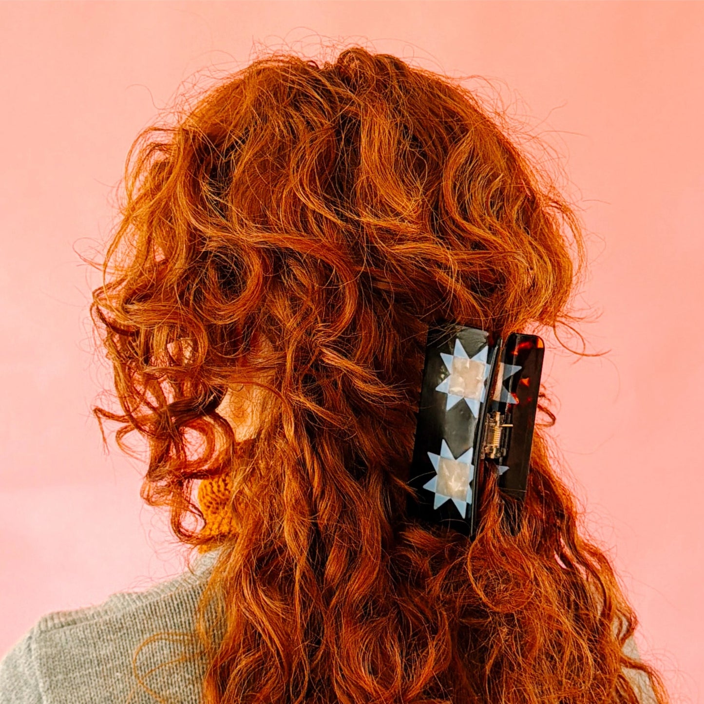 A close-up of a person with long, curly red hair wearing a black claw clip decorated with white star patterns, set against a solid pink background.