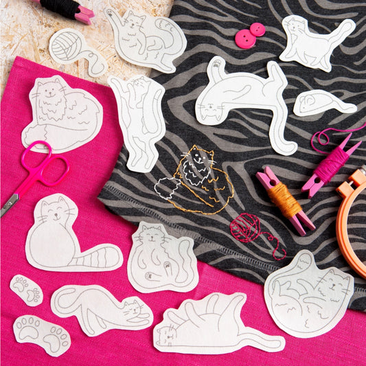 A flat-lay of cat-shaped fabric cut templates or appliqués scattered across bright pink and black-and-white zebra-patterned fabric, with embroidery floss, buttons, scissors, and an embroidery hoop nearby.