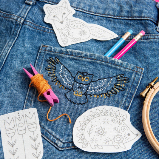 A styled flat-lay of a denim pocket with embroidered owl artwork, alongside matching white paper templates featuring a fox and a hedgehog, plus embroidery floss, pencils, and an embroidery hoop on a blue denim background.
