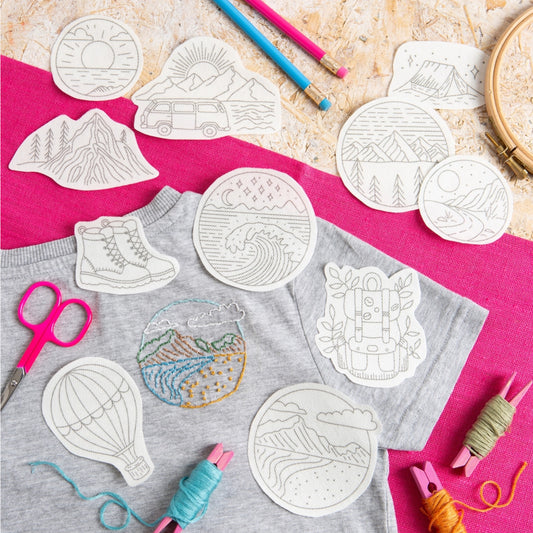 A styled flat-lay of a gray t-shirt with several white circular and shaped embroidery templates placed on top, alongside craft supplies including pink scissors, colored pencils, thread spools, and a wooden hoop on a bright pink and beige background.