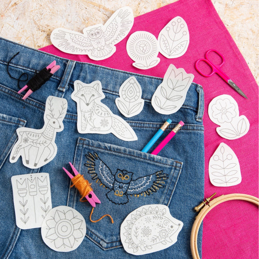 A styled flat-lay of embroidered animal and floral design templates placed on blue denim jeans, with a bright pink fabric background, scissors, thread, pencils, and an embroidery hoop.