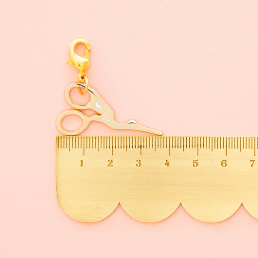 Gold charm shaped like a pair of scissors with a scalloped edge ruler on a pink background