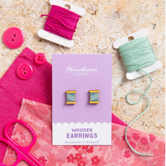 Wooden earrings on a Hawthorn card with pink and green thread spools and buttons on a textured surface.