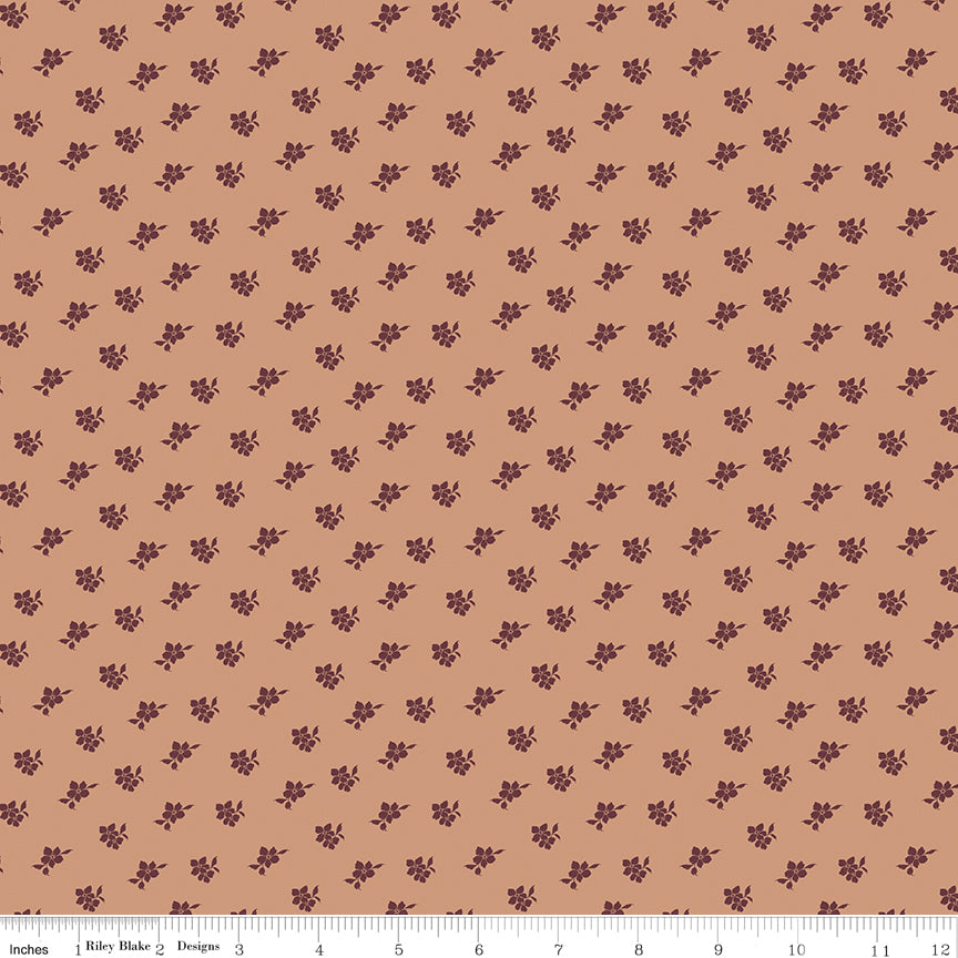 A repeating pattern of small dark brown floral motifs on a warm tan background, shown as a fabric or paper swatch with a ruler along the bottom edge.