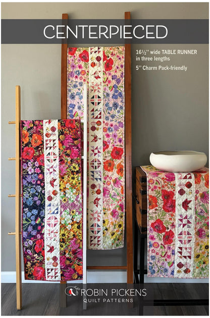 Promotional image for a quilt pattern titled 'CENTERPIECED' by Robin Pickens Quilt Patterns, showing three colorful floral table runners displayed on wooden stands in an indoor room setting.