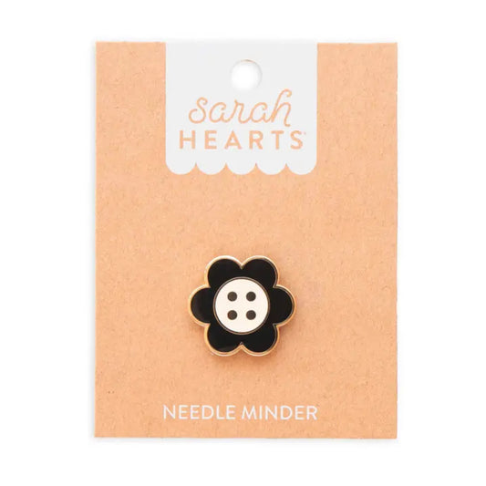 A flower-shaped enamel needle minder with a black petal and a white center featuring four black dots, displayed on a peach-colored card labeled 'sarah HEARTS' and 'NEEDLE MINDER'.