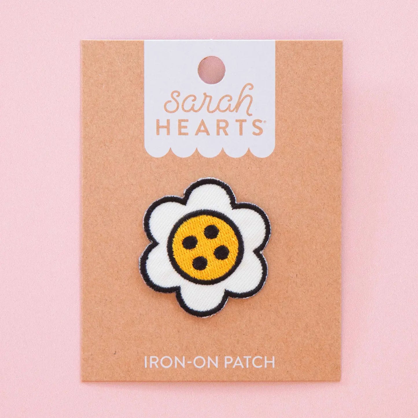 A daisy-shaped iron-on patch with white petals, a black stitched outline, and a yellow center with black dots, displayed on a kraft paper backing card labeled 'Sarah Hearts' against a soft pink background.