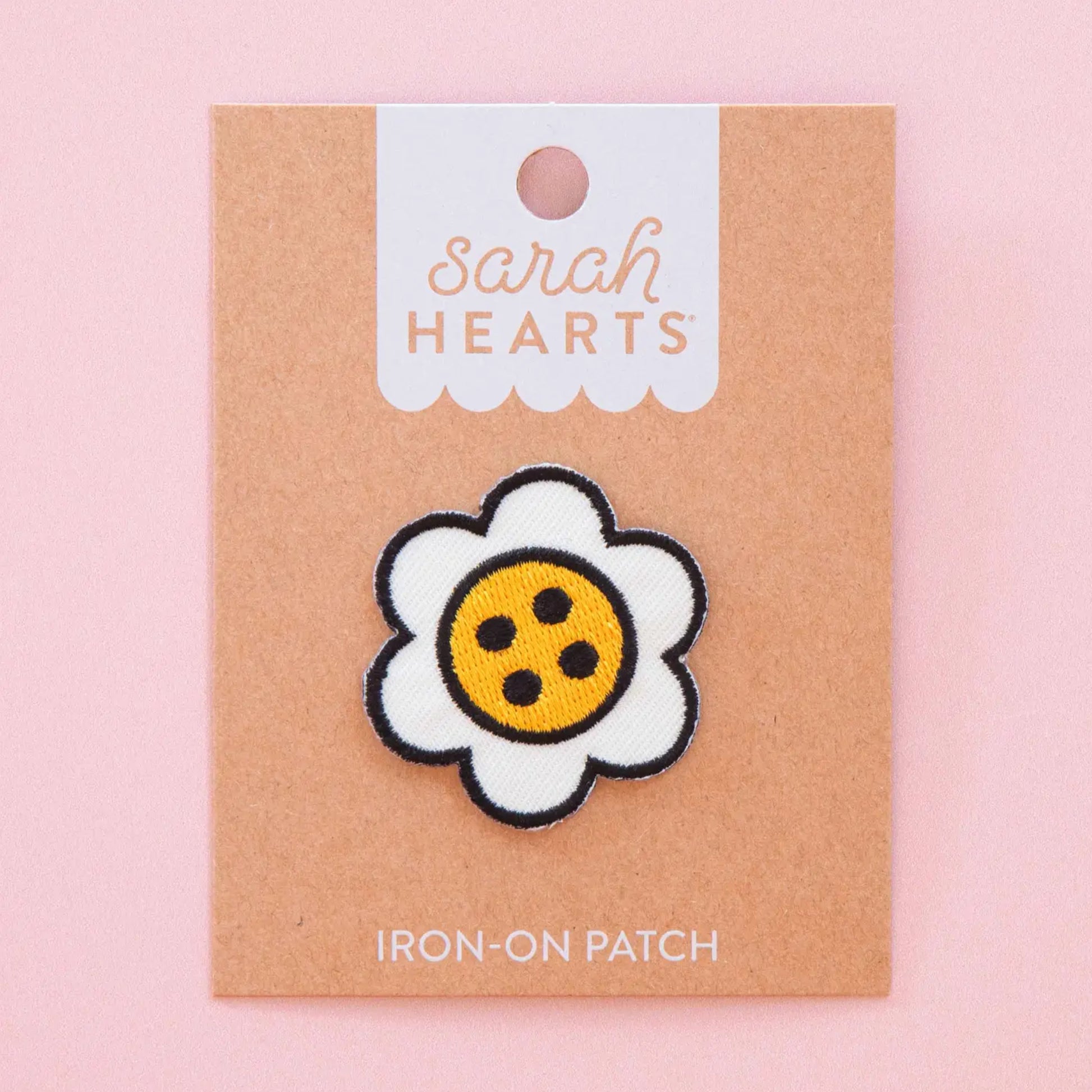 A daisy-shaped iron-on patch with white petals, a black stitched outline, and a yellow center with black dots, displayed on a kraft paper backing card labeled 'Sarah Hearts' against a soft pink background.