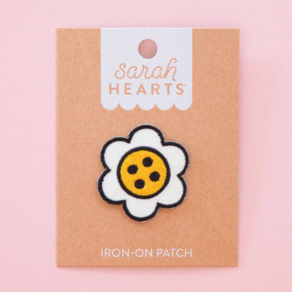 A daisy-shaped iron-on patch with white petals, a black stitched outline, and a yellow center with black dots, displayed on a kraft paper backing card labeled 'Sarah Hearts' against a soft pink background.