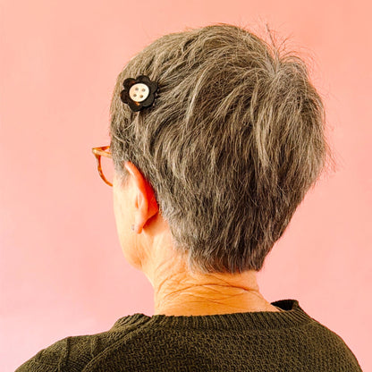 A close-up back view of a person with short gray hair wearing a black flower-shaped hair clip with a white button, set against a solid pink background.