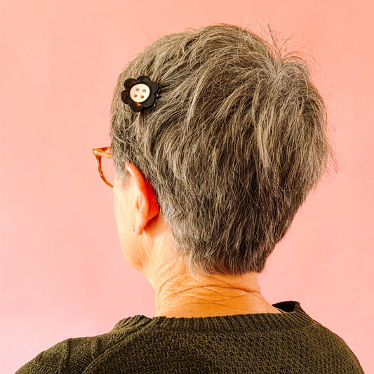 A close-up back view of a person with short gray hair wearing a black flower-shaped hair clip with a white button, set against a solid pink background.