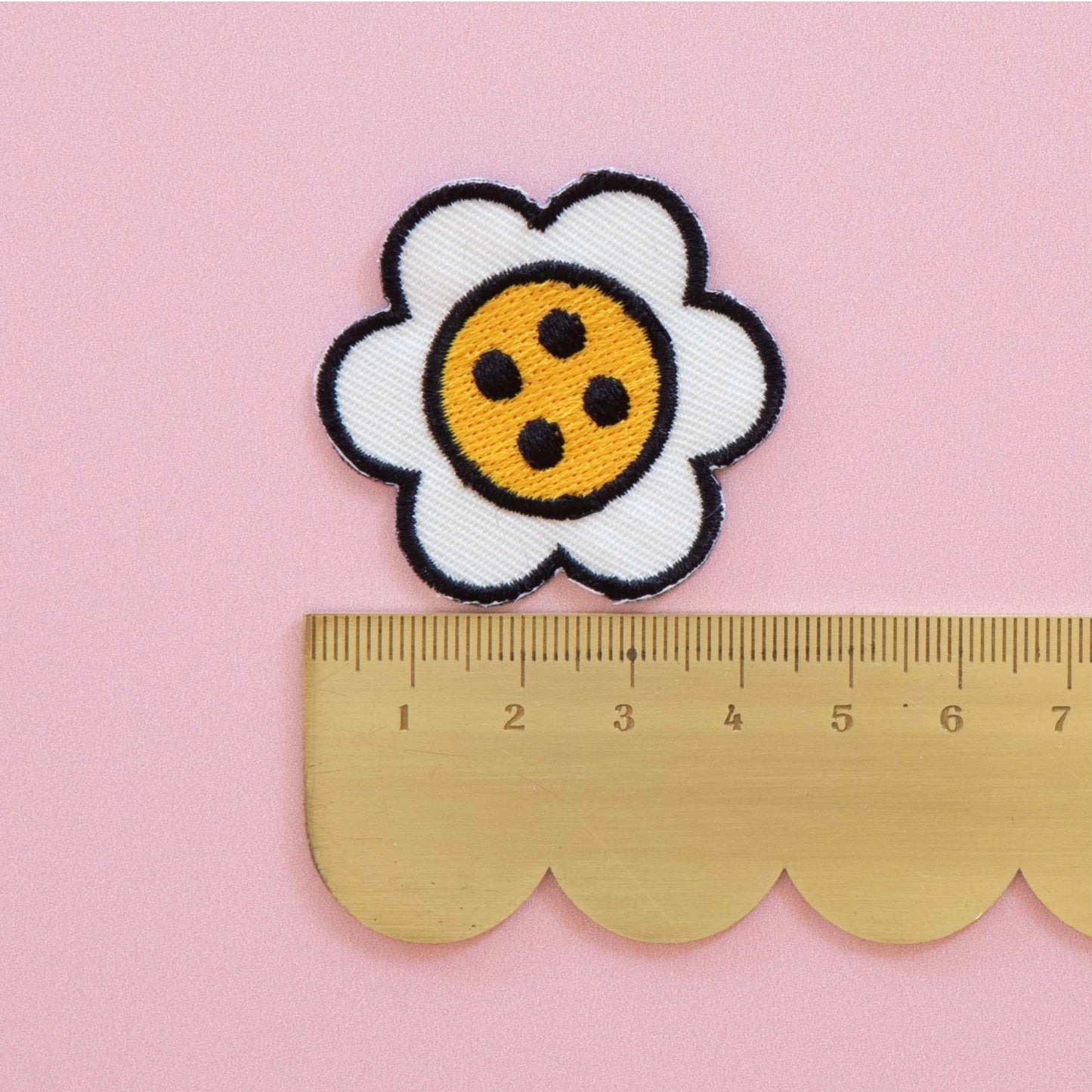 A white daisy patch with a yellow center and black outlines, shown above a gold metal ruler on a pink background to indicate size.