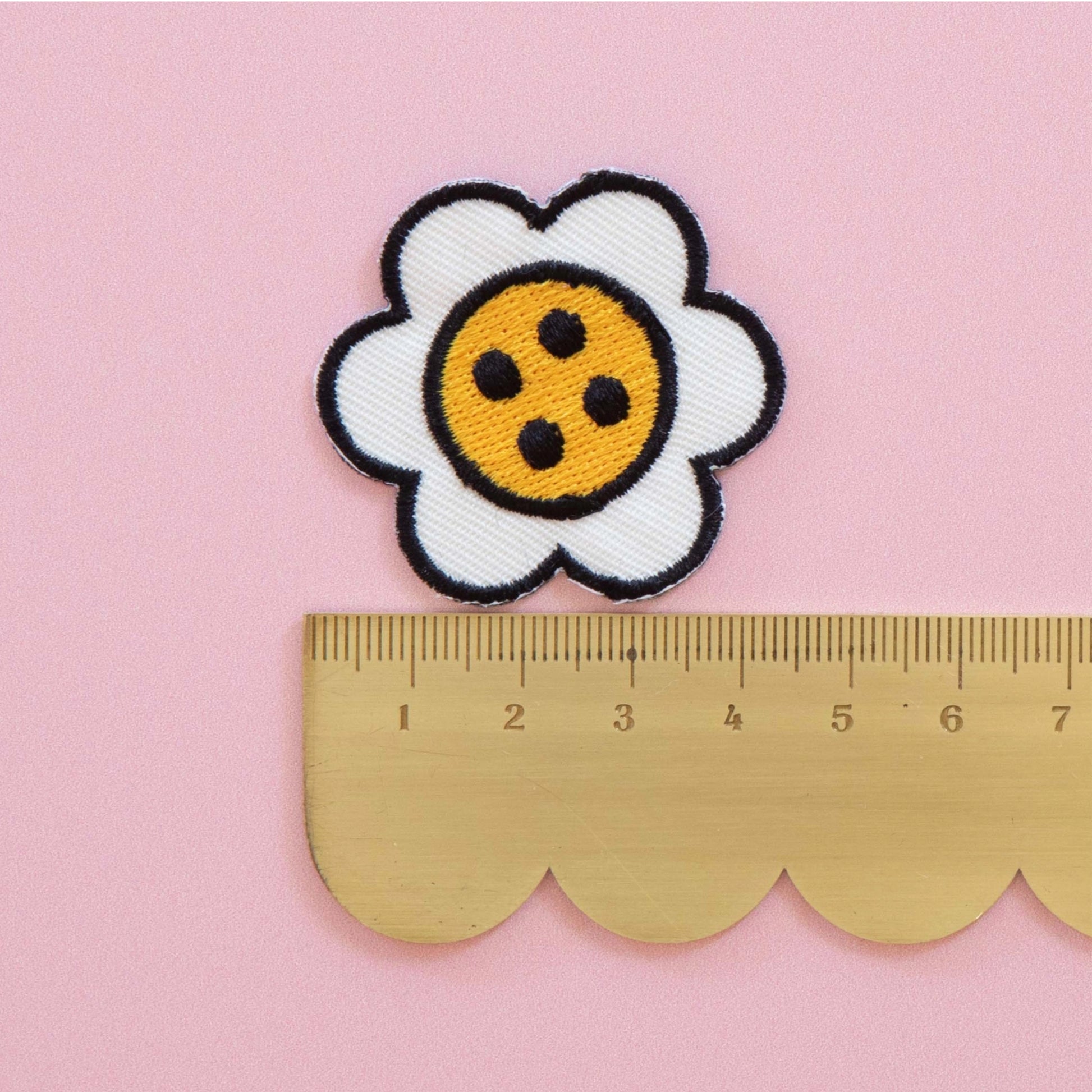 A white daisy patch with a yellow center and black outlines, shown above a gold metal ruler on a pink background to indicate size.
