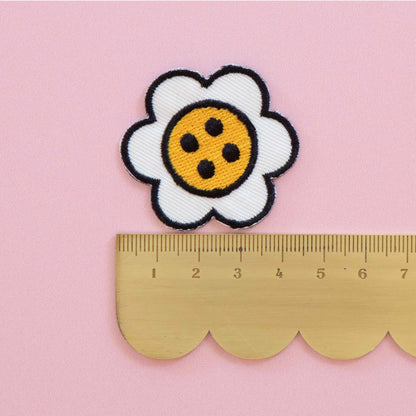 A white daisy patch with a yellow center and black outlines, shown above a gold metal ruler on a pink background to indicate size.
