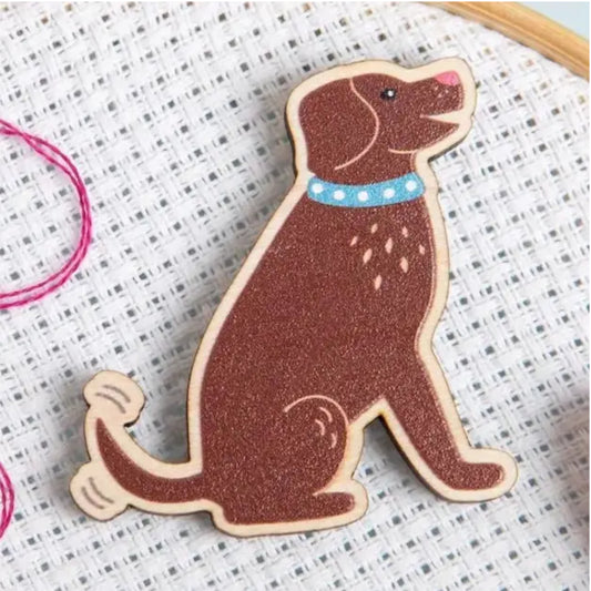 A wooden dog-shaped needle minder with a brown finish, featuring a light blue collar with white polka dots, displayed on a white cross-stitch fabric background.