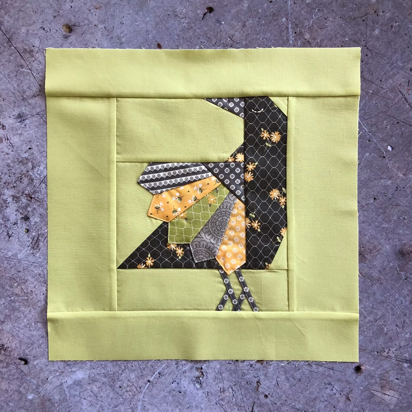 A square quilted fabric block featuring a stylized bird made from multiple patterned fabrics in yellow, orange, green, gray, and black, set against a solid pale yellow border, displayed on a textured gray surface.