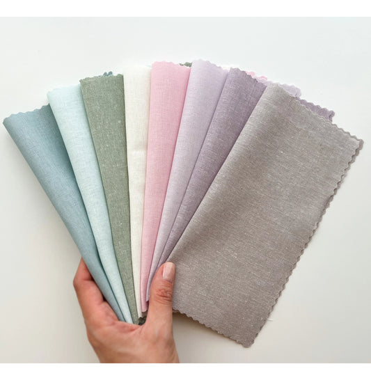 A hand holds a fan of eight fabric swatches in soft pastel and neutral shades, including light blue, pale green, off-white, pink, lavender, lilac, and taupe, against a plain white background.