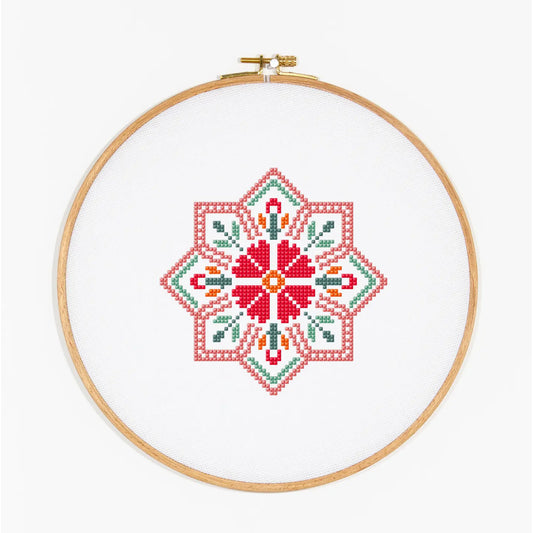A completed cross-stitch design featuring a geometric floral pattern in red, green, orange, and white, held in a wooden embroidery hoop against a plain white background.