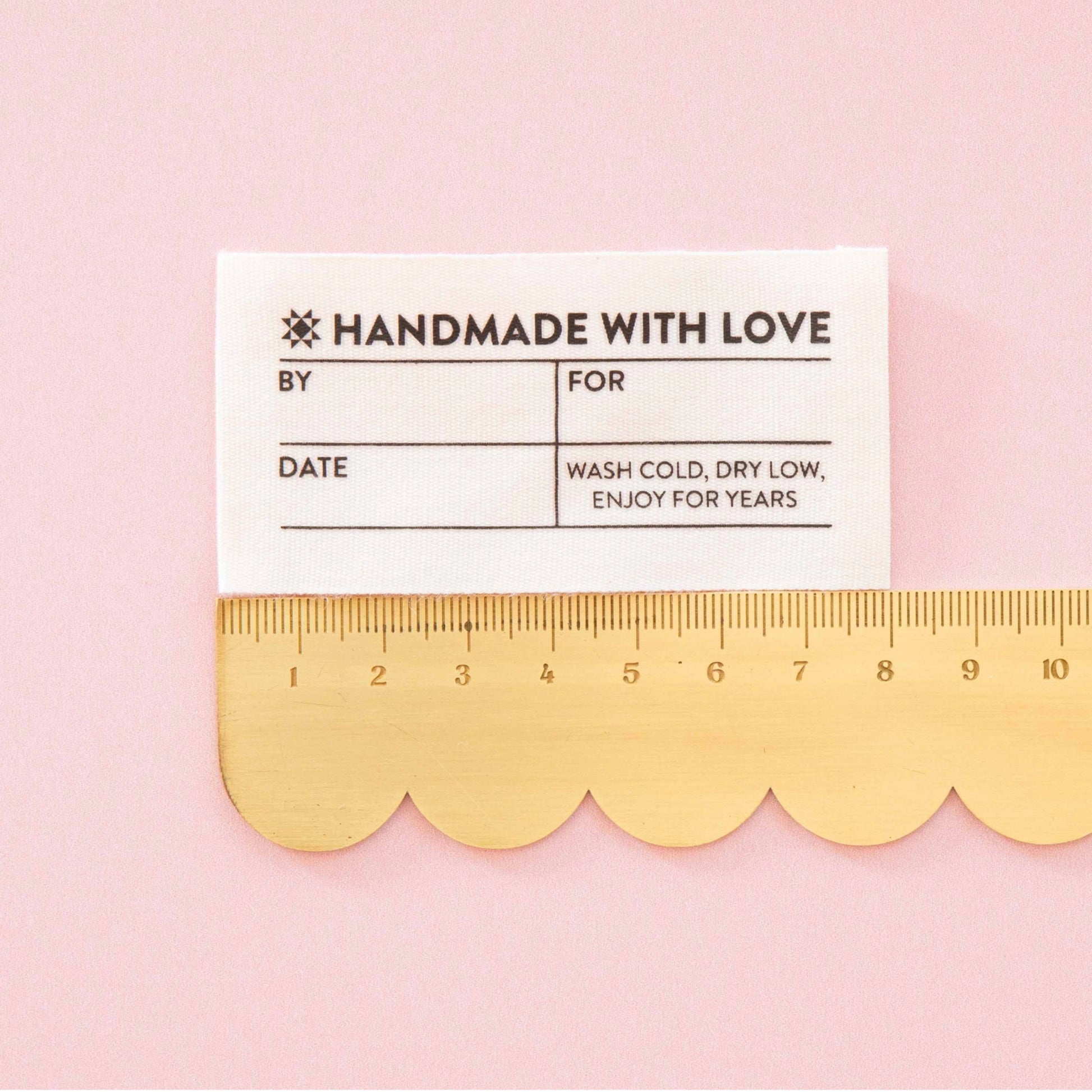 A white rectangular fabric tag with black printed text reading 'HANDMADE WITH LOVE' and care instructions, placed above a gold scalloped ruler on a light pink background.