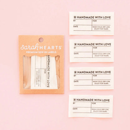 Sarah Hearts premium sew-in labels shown in a kraft paper sleeve on a light pink background, alongside four individual cream labels with printed fields and decorative text.