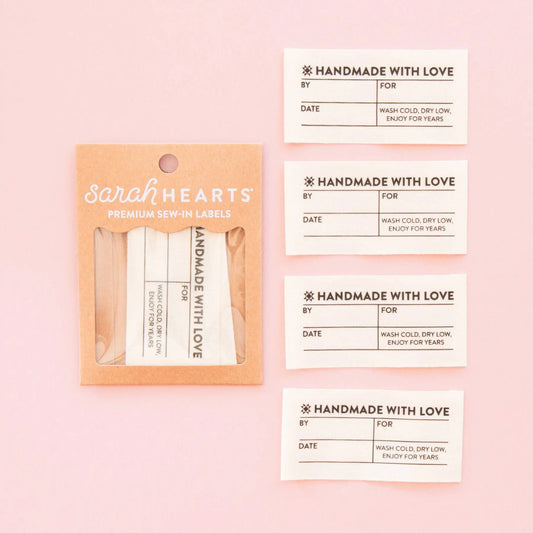 Sarah Hearts premium sew-in labels shown in a kraft paper sleeve on a light pink background, alongside four individual cream labels with printed fields and decorative text.