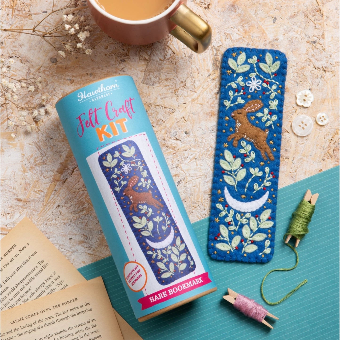 A felt craft kit for making a hare bookmark, shown with its light blue cylindrical packaging and the finished blue embroidered hare bookmark placed beside it on a teal mat. The scene includes a mug, book pages, buttons, and thread spools on a textured beige surface.