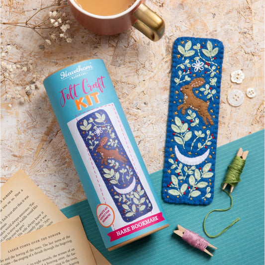 A felt craft kit for making a hare bookmark, shown with its light blue cylindrical packaging and the finished blue embroidered hare bookmark placed beside it on a teal mat. The scene includes a mug, book pages, buttons, and thread spools on a textured beige surface.
