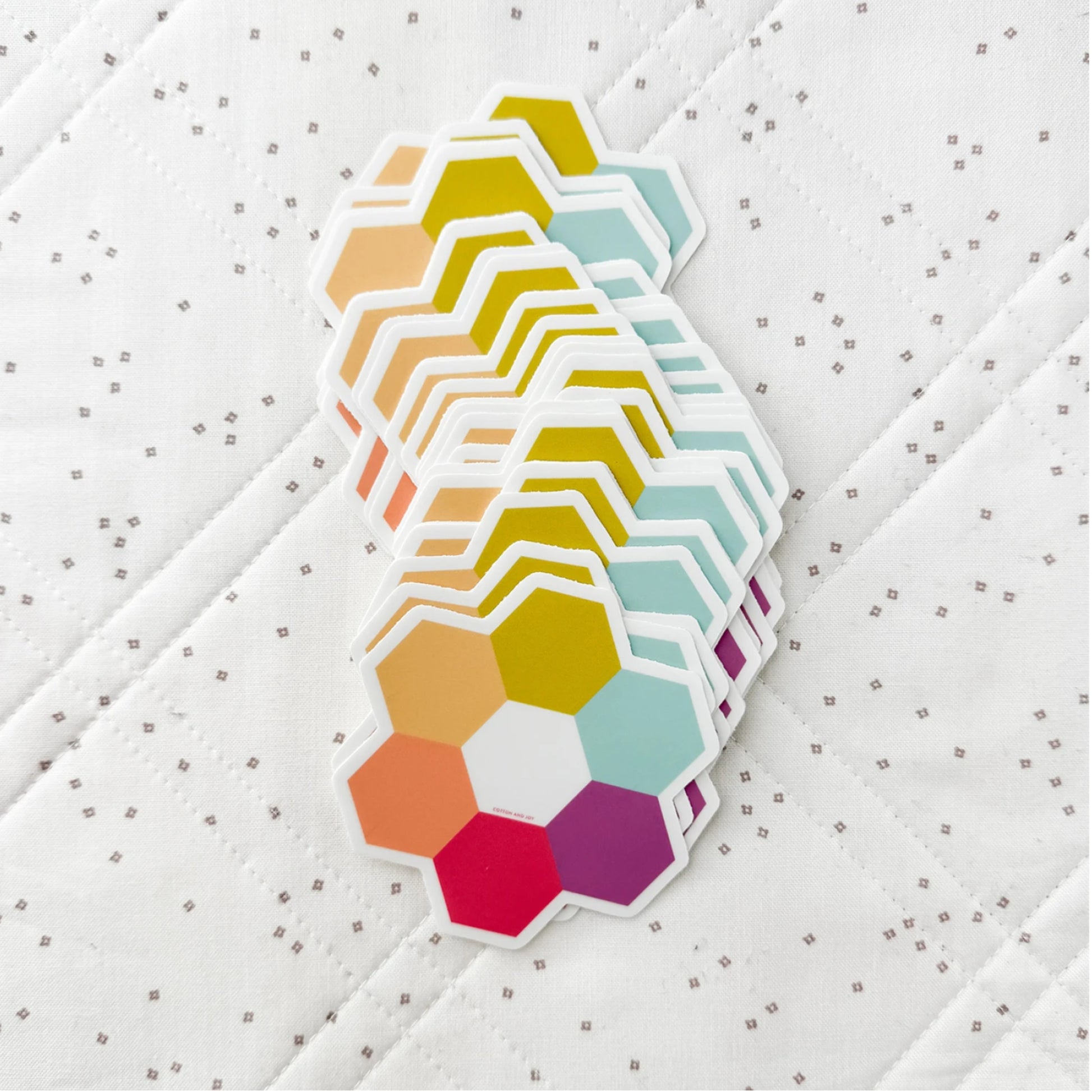 A stack of hexagonal sticker or patch pieces in assorted pastel and bright colors, including yellow, orange, red, purple, and mint, arranged on a white quilted fabric background.