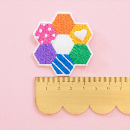 A colorful embroidered patch shaped like a flower with six hexagonal sections in pink, orange, yellow, purple, blue, and green, shown above a wooden ruler on a light pink background.