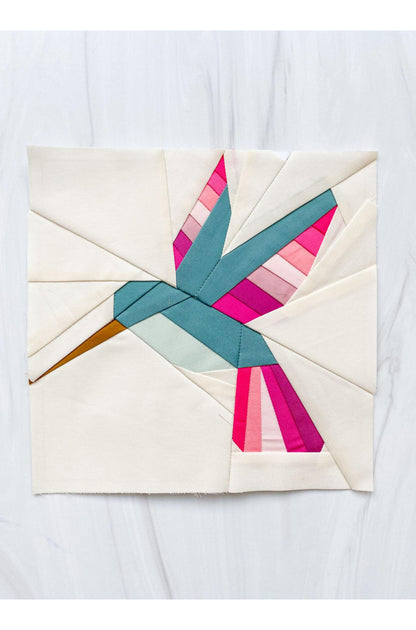 A square fabric art piece featuring a geometric hummingbird design in teal, pink, magenta, and orange on an off-white background, displayed flat on a light gray marble surface.