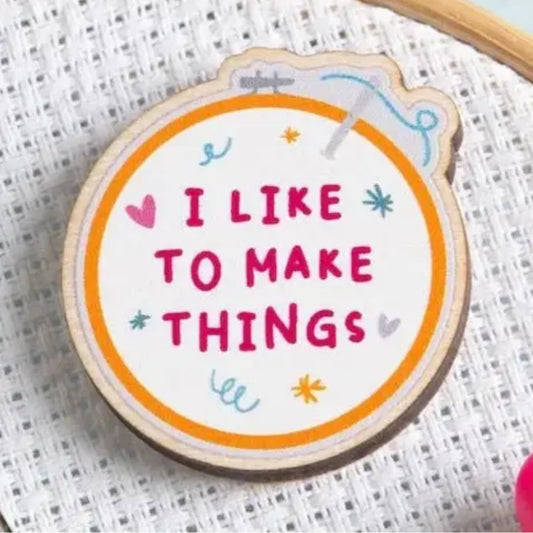 A round wooden embroidery hoop needle minder with the phrase 'I LIKE TO MAKE THINGS' stitched in pink letters, surrounded by small colorful decorative motifs on a white textured fabric background.