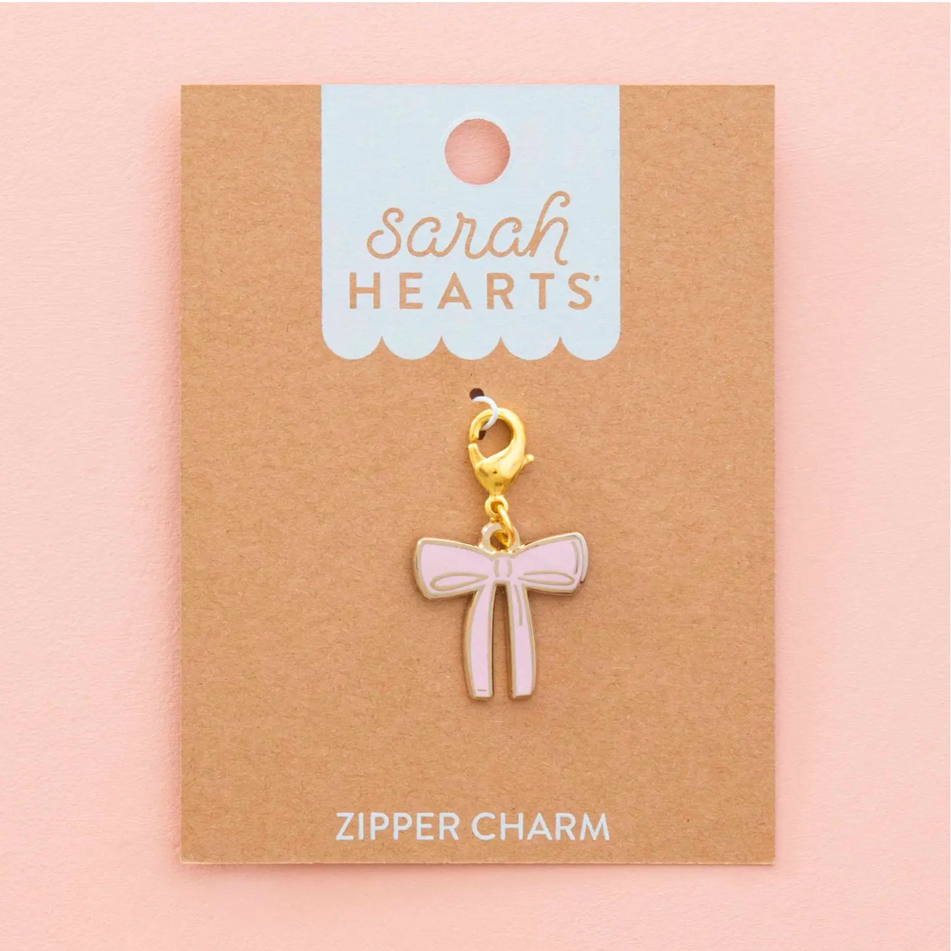 A pink bow-shaped zipper charm with a gold clasp, displayed on a brown kraft card labeled 'Sarah Hearts' against a soft pink background.