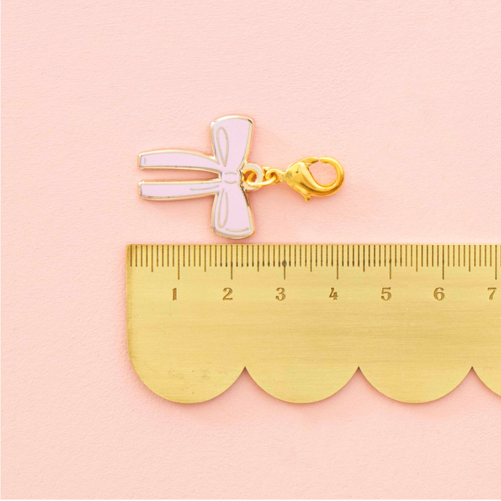A small bow-shaped charm or pendant in pale pink enamel with a gold clasp, placed above a gold ruler on a soft pink background.