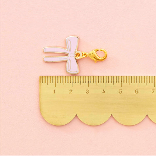 A small bow-shaped charm or pendant in pale pink enamel with a gold clasp, placed above a gold ruler on a soft pink background.