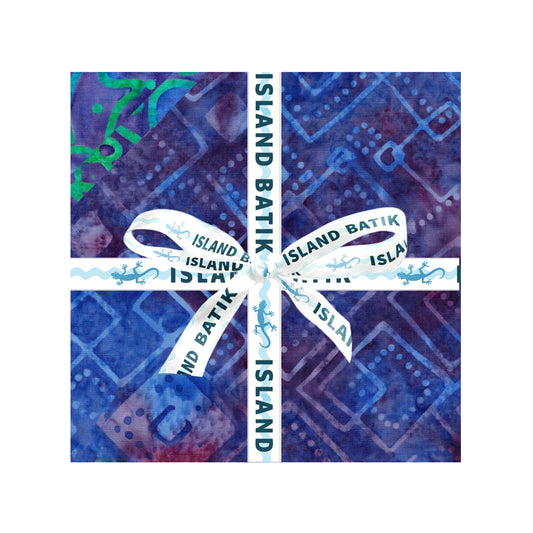 A square fabric bundle in blue and purple batik-patterned paper, tied with a white ribbon printed with 'ISLAND BATIK' and small lizard motifs, centered on a white background