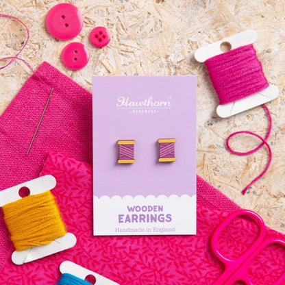 Wooden earrings on a purple card with pink and yellow thread spools and buttons on a textured surface.
