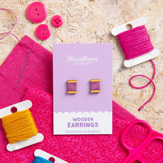Wooden earrings on a purple card with pink and yellow thread spools and buttons on a textured surface.