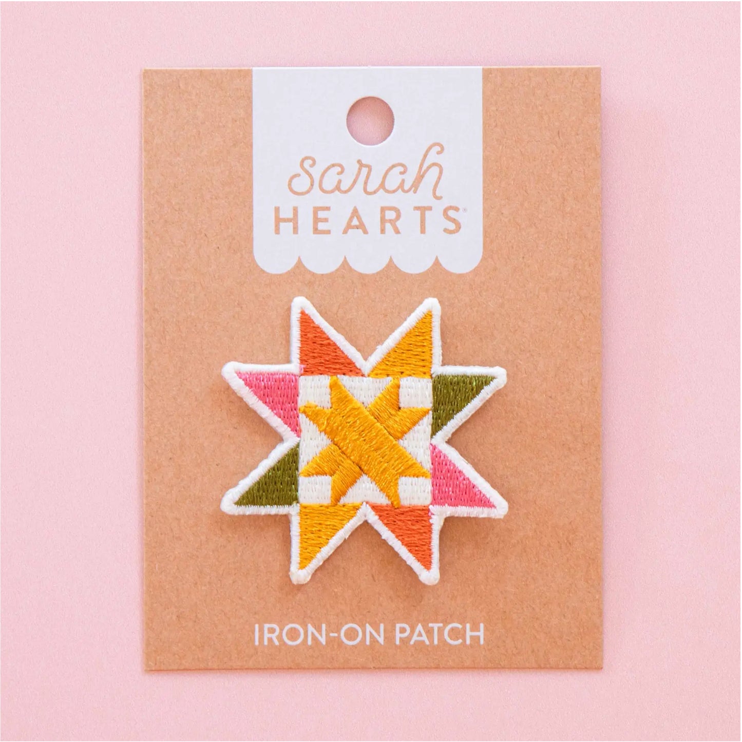 A star-shaped embroidered iron-on patch by Sarah Hearts, featuring a geometric quilted pattern in orange, yellow, pink, olive green, and white, mounted on a kraft paper backing card against a solid pink background.