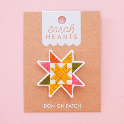 A star-shaped embroidered iron-on patch by Sarah Hearts, featuring a geometric quilted pattern in orange, yellow, pink, olive green, and white, mounted on a kraft paper backing card against a solid pink background.