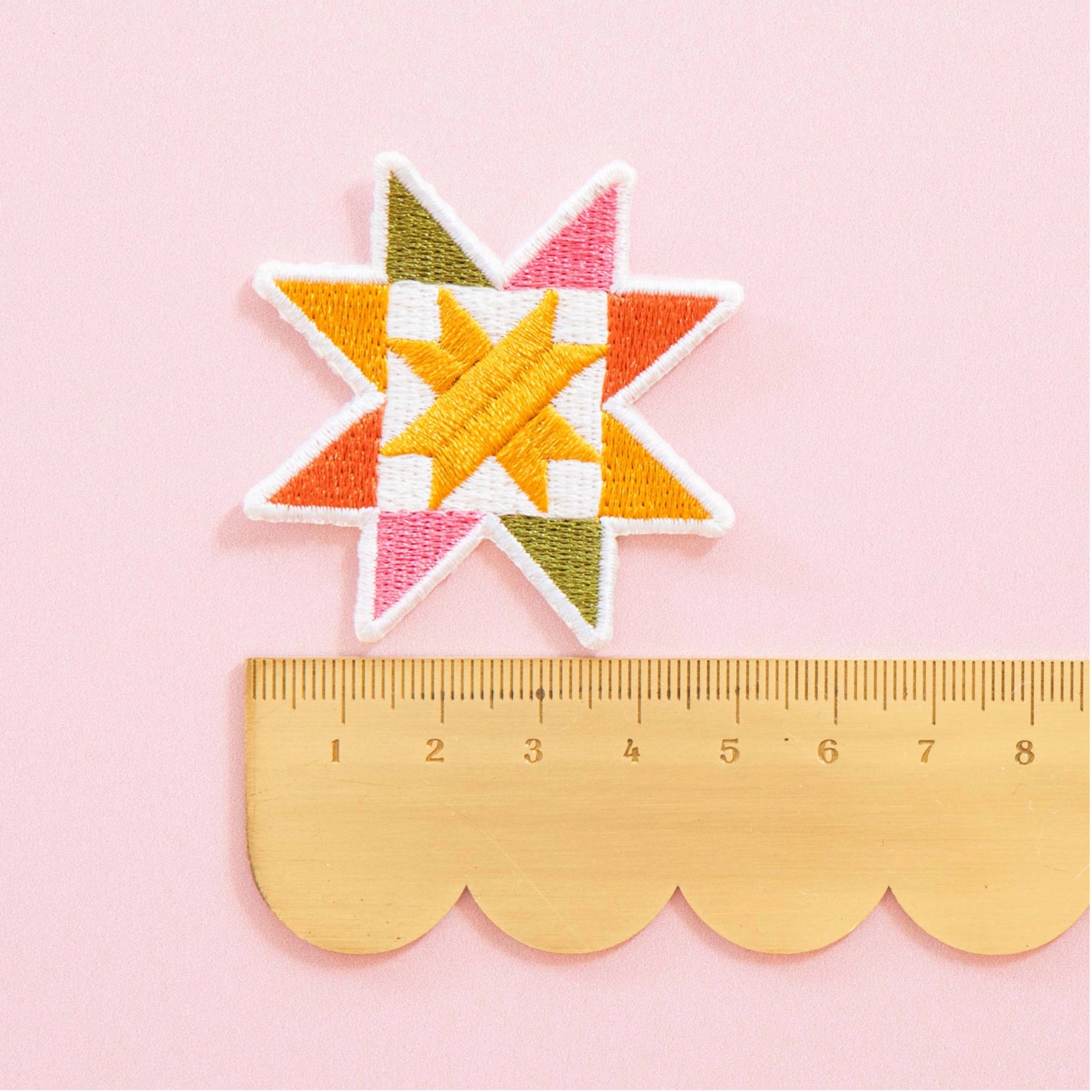 A colorful embroidered star patch with a white border, shown above a wooden ruler on a light pink background. The patch features geometric star-like shapes in orange, yellow, pink, olive green, and white.