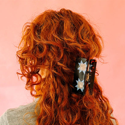 A close-up of a person with long, curly red hair wearing a black claw clip decorated with white star patterns, set against a solid pink background.