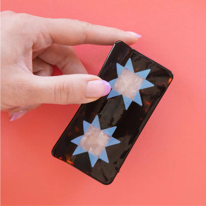 A hand holds a glossy hair claw with two blue six-pointed star cutouts over pale pink square inserts, set against a solid coral-pink background.