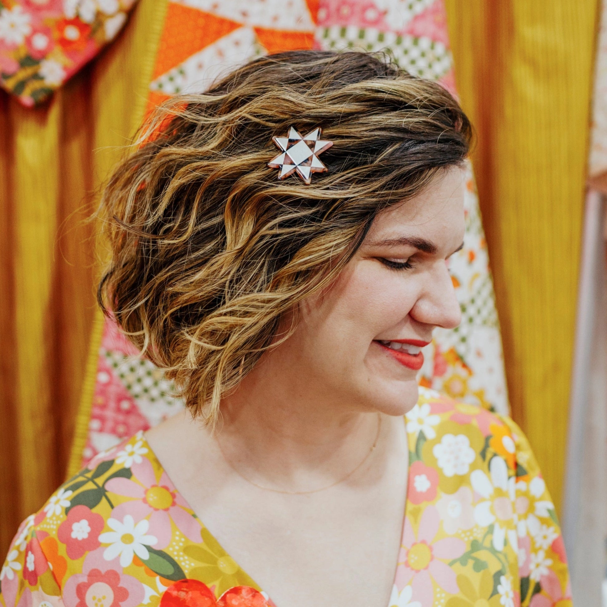 Woman with a decorative hair clip in her hair, wearing a colorful floral outfit against a patterned background.