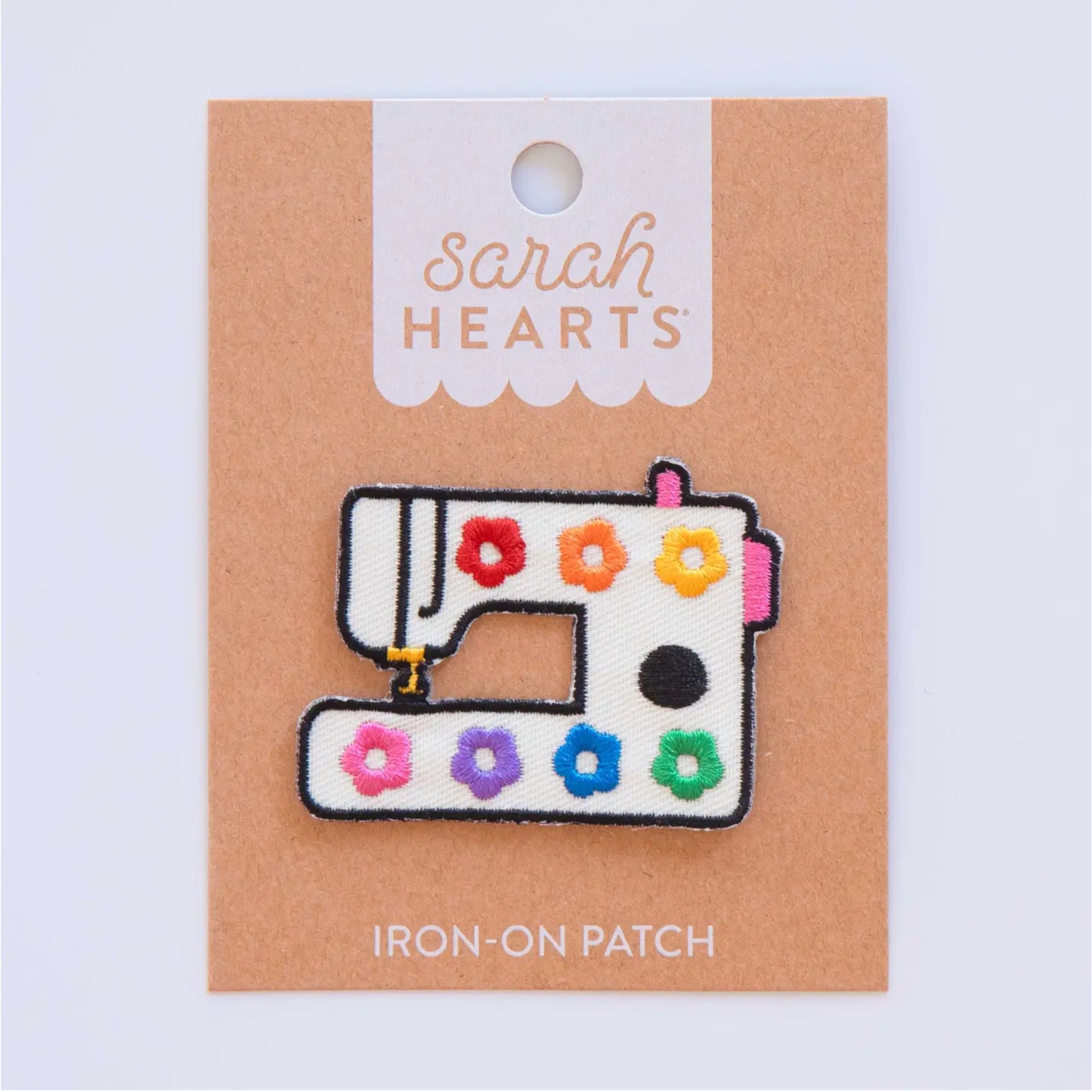 A sewing machine iron-on patch by Sarah Hearts, featuring a white fabric base decorated with colorful embroidered flowers in red, orange, yellow, pink, purple, blue, and green, mounted on a brown kraft backing card.